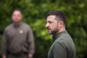 Володимир Зеленський у профіль на тлі зеленого листя, позаду розмитий чоловік