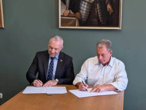 Двоє чоловіків підписують документи за столом на тлі історичної картини