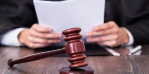 Деревяний судовий молоток на столі, за ним людина в костюмі читає папери